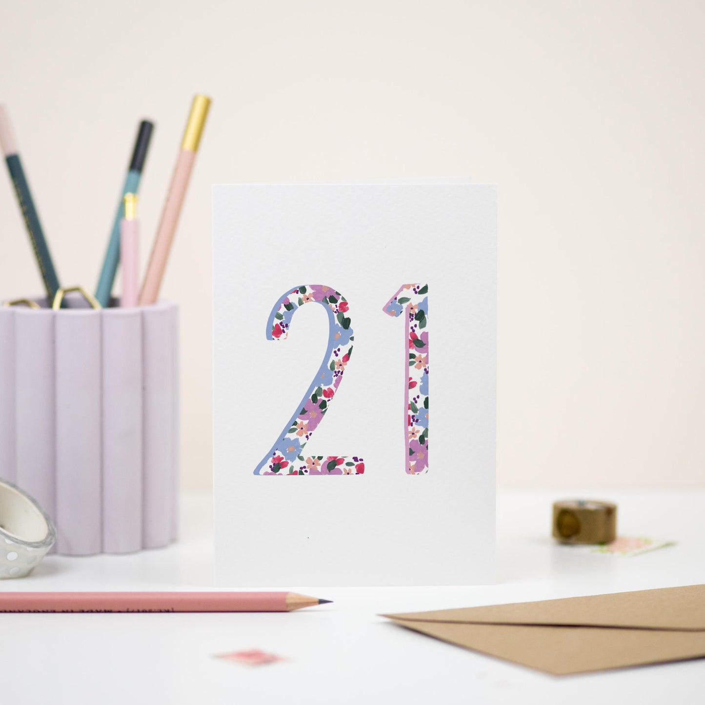 A floral 21st birthday card on a white background card. The card is displayed  with a pencil holder in the background and a pencil in the foreground.