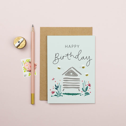 A beehive in the middle of the card surrounded by flowers and bees on either side. The lettering reads Happy Birthday. 